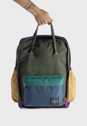 Mochila colorida hecha de tela resistente. Cuenta con un cuerpo principal verde, bolsillo delantero azul y verde azulado, y bolsillos laterales de color beige. Incluye asas robustas.