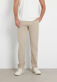 Pantaloni beige a gamba dritta realizzati in cotone con una texture liscia. Abbinati a sneakers bianche e una camicia bianca aderente. Nessun motivo visibile.