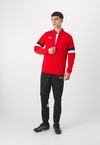 Rød sportjakke med svart og hvite striper, glidelås foran, kombinert med svarte treningsbukser med hvit aksent. Svarte joggesko.
