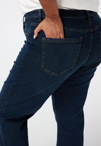 Donkerblauwe denim jeans met een aangesloten ontwerp, voorzien van een achterzak en zichtbare stiksels. Het materiaal vertoont een gladde textuur.