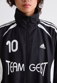 Černá sportovní bunda s bílými akcenty, má vysoký límec, zavírání na zip, velké číslo "10" a nápis "TEAM GEIST" vytištěný na přední straně, značkovaná logem Adidas.