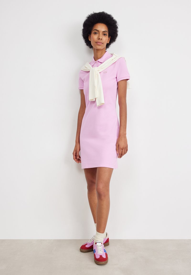 Vestido polo rosa con mangas cortas, con un suéter de punto blanco atado alrededor de los hombros y zapatillas deportivas multicolores.