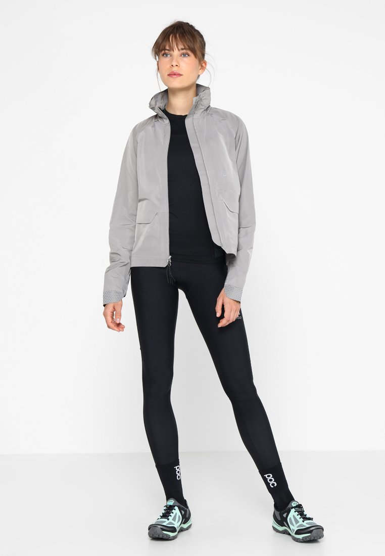 Hellgraue Jacke mit Stehkragen, zwei vorderen Taschen und strukturierten Bündchen, kombiniert mit schwarzen Leggings und schwarzen Sportschuhen mit mintfarbenen Akzenten.