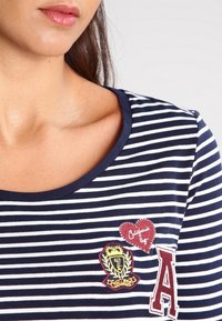 Marinblå och vit randig T-shirt med rundad halsringning, prydd med broderade lappar: ett hjärta och ett märke med siffran 7 och bokstaven A.