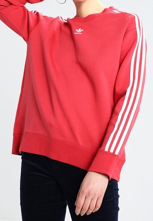 Osoba ubrana w czerwoną bluzę Adidas z białymi paskami na rękawach i logo na piersi, w połączeniu z ciemnymi spodniami.