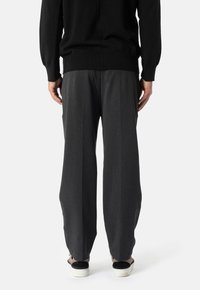 Šedé, skládací kalhoty s uvolněným střihem vyrobené z texturované látky. V kombinaci s černými nazouvacími botami, pohled zezadu.