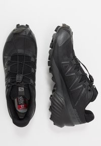 Chaussures de sport noires avec une surface texturée, tige en mesh et semelle en caoutchouc agressive. Dotées de lacets élastiques et d'éléments de rembourrage.