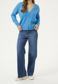 Modrý pletený kardigan s výstřihem do V a pěti knoflíky, kombinovaný s tmavomodrými širokými džíny. Na nohou jsou robustní bílé tenisky.