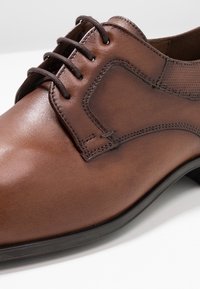 Brune læderdress sko med en glat finish, der har et snørebåndsdesign og syninger. Sålen er svagt buet for støtte.