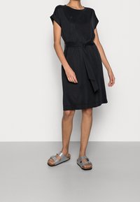 Schwarzes kurzärmeliges Kleid mit einem taillierten Gürtel, leicht lockerer Passform und glatter Textur. Kombiniert mit grauen Sandalen mit einer Schnallendetails.