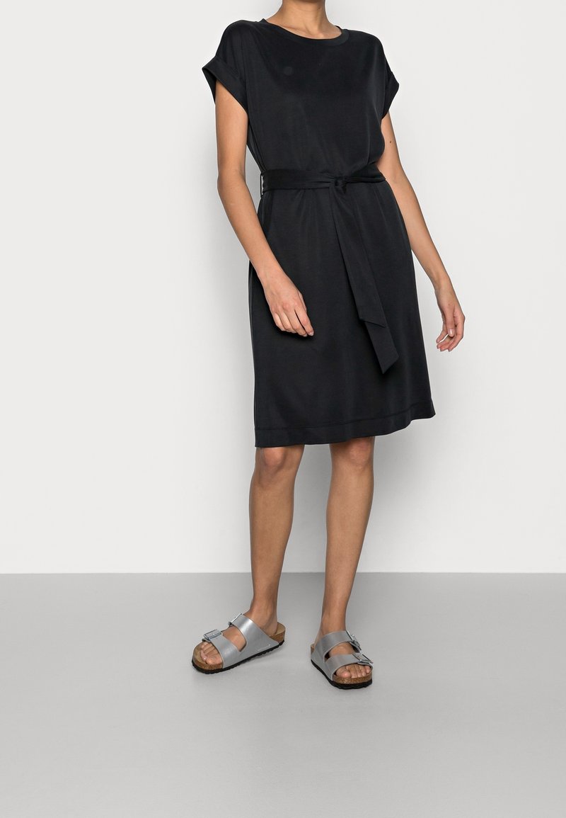 Schwarzes kurzärmeliges Kleid mit einem taillierten Gürtel, leicht lockerer Passform und glatter Textur. Kombiniert mit grauen Sandalen mit einer Schnallendetails.