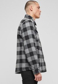 Pilka ir juoda languota flanelinė striukė ilgomis rankovėmis, su sagų užsegamais rankogalais ir tiesiu kirpimu. Yra matomos šoninės kišenės ir apykaklė.