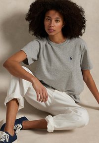 Kvinne med krøllete hår iført en grå t-skjorte, hvite oppkrempede bukser og navy sneakers som sitter på gulvet mot en beige bakgrunn.
