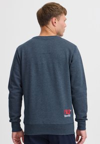 Sudadera gris oscuro hecha de tejido texturizado, con cuello redondo y mangas largas. Logo rojo con "Desde 1993" en la parte inferior derecha.