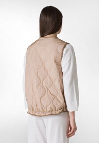 Beige gesteppte Weste mit geripptem Saum und einem strukturierten Muster. Wird über einem weißen Langarmshirt getragen und zeigt ein einfaches, klares Design.