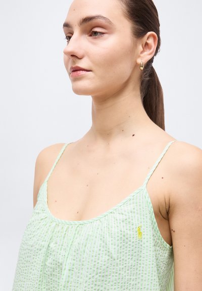 Νεαρή γυναίκα με αμάνικο τοπ με ανοιχτοπράσινες ρίγες, χρυσό σκουλαρίκι σε σχήμα δάκρυ, και μαλλιά δεμένα πίσω, κοιτάζοντας πλάγια μπροστά σε απλό φόντο.