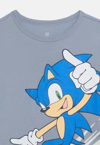 Μπλε καρτούν σκαντζόχοιρος με πράσινα μάτια και λευκά γάντια που δείχνει προς τα πάνω σε γκρι παιδικό γιακά μπλουζάκι με ετικέτα GAP KIDS.