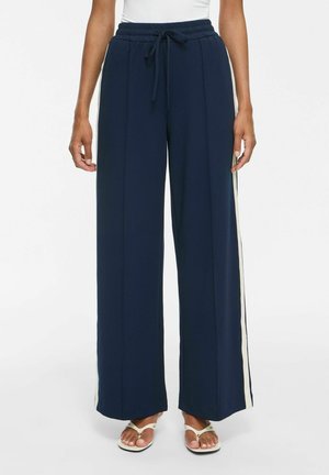 Pantalon large bleu marine avec bandes blanches sur les côtés, taille élastique avec cordon, porté avec des sandales blanches et un haut blanc.