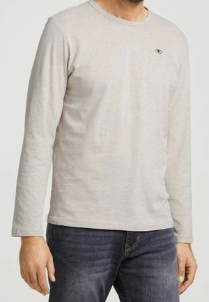 Homme portant une chemise beige clair à manches longues avec un petit logo sur la poitrine et un jean bleu foncé.