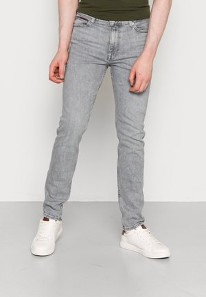 Mężczyzna w jasnoszarych dopasowanych jeansach, białych trampkach i oliwkowej koszuli, stojący przy gładkiej ścianie.