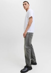 Tânăr stând deoparte, îmbrăcat cu un tricou alb, blugi gri și pantofi negri cu șireturi, pe un fundal luminos uniform.