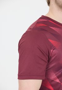Mand iført en maroon kortærmet sportsbluse med røde og mørke geometriske mønstre, beskåret for at vise skulder og arm.