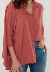 Frau trägt eine lockere korallrosa Bluse mit Dreiviertelärmeln, geschichtete goldene Halsketten und blaue, verwaschene Jeans, mit der Hand in der Tasche.