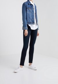 Giacca di denim blu, con cuciture visibili e due tasche sul petto. Abbinata a jeans skinny scuri e sneaker bianche. Texture lisce in tutto.