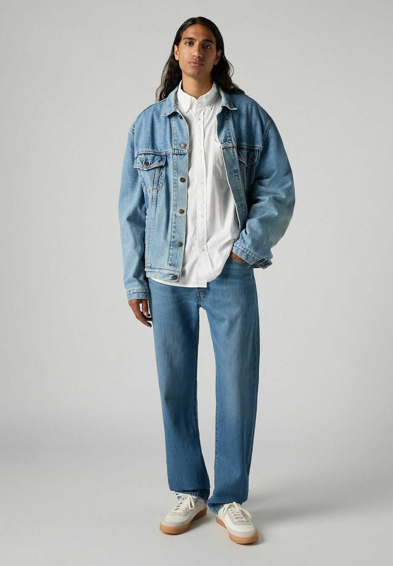 Veste en denim et jean bleu clair associés à une chemise blanche à boutons et des baskets beiges. Coupe décontractée notable et couleur délavée.