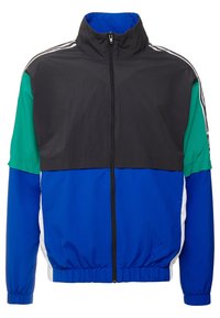 Färgglad träningsjacka med en svart överdel, gröna ärmar, blå nederdel, elastiska mudd och hög krage med dragkedja.