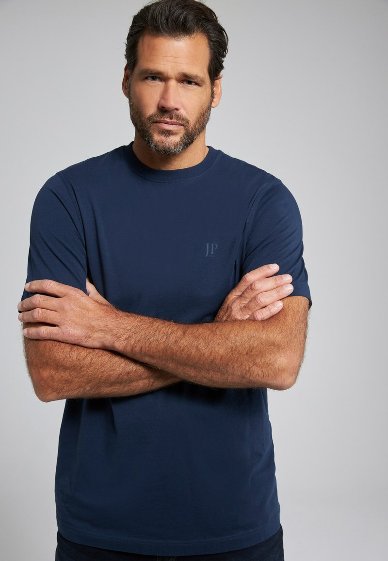 Camiseta de manga corta azul marino hecha de una mezcla de algodón, con un pequeño logo en el pecho. El modelo tiene los brazos cruzados y una expresión neutral.