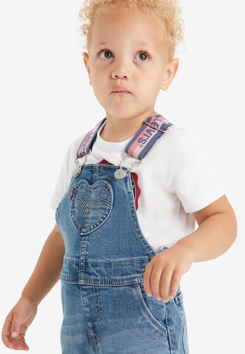 Jeans-Latzhose mit einer herzförmigen Tasche, ausgestattet mit verstellbaren rosa und blauen Trägern. Getragen über einem weißen Shirt mit einem roten Logo-Detail.