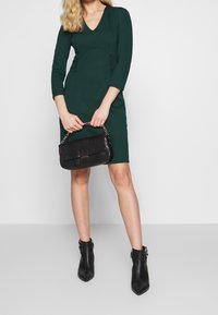 Green fitted dress with a V-neck, three-quarter sleeves, and ruched sides. Black textured handbag with gold hardware and chain strap. Black ankle boots.