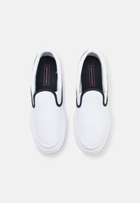 Ein Paar weiße Slip-On-Sneaker mit schwarzem Rand und Tommy Hilfiger-Logo auf schwarzen Innensohlen, von oben auf einem weißen Hintergrund betrachtet.