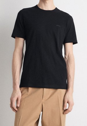 Mand iført en simpel sort kortærmet T-shirt med subtilt "BOSS"-logo og beige bukser, stående mod en lys baggrund.