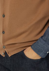 Rudas raukšlėtas kardiganas su juodais mygtukais, dėvimas ant mėlyno šambro marškinėlių. Tamsiai mėlynos džinsinės kelnės su matomu priekinio kišenės.