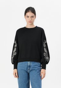 Svart hettegenser med lange ermer som har intrikate hvite blomsterbroderier på ermene. Ribbet nederkant og rund halslinning. Blå jeans brukt.