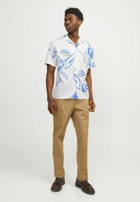 Camisa de manga corta, blanca, con estampados florales azules y acentos marrones, combinada con pantalones color beige y zapatos oscuros. Tiene un acabado texturizado.