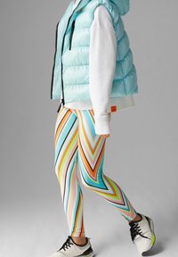 Světle modrá vatovaná vesta přes bílou mikinu, doplněná barevnými legínami se vzorem cikcak v oranžových, růžových a zelených pruzích. Bílé tenisky.