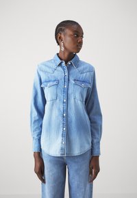 Chemise en denim bleu clair avec un bouton sur le devant, deux poches poitrine et des manches longues. Présente des coutures contrastantes et un ourlet arrondi.