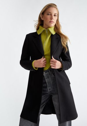 Cappotto nero con un design sartoriale, maglione verde brillante in contrasto sotto, e jeans in denim scuro con una leggera lucentezza.
