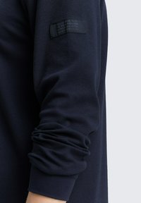 Marineblauwe langemouwtop met een gestructureerde afwerking, voorzien van een logopatch op de mouw. De manchetten zijn iets gerimpeld voor een ontspannen pasvorm.