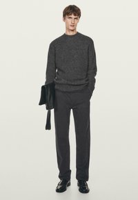 Ciemnoszary sweter dzianinowy z okrągłym dekoltem, zestawiony z ciemnoszarymi spodniami. Model trzyma czarną skórzaną torbę i ma na sobie czarne buty.