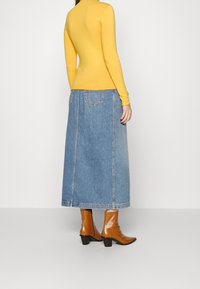 Maglia gialla a maniche lunghe abbinata a una gonna lunga in denim blu, con due tasche posteriori e stivali con tacco marroni. Texture lisce in tutto.