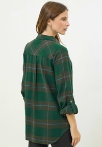 Camicia a scacchi verde con colletto, con maniche arrotolate e vestibilità morbida. Motivi in verde scuro, marrone e blu. Texture del tessuto morbida.