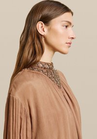 Abito marrone a pieghe con colletto alto caratterizzato da intricate perline. Tessuto liscio con una leggera lucentezza, impreziosito da delicati dettagli attorno al collo.