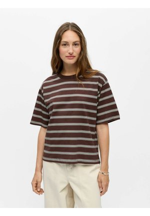 Femme aux longs cheveux bruns portant un t-shirt à manches courtes rayé marron et blanc et un pantalon beige clair sur un fond blanc uni.