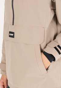 Vedenkestävä beigen värinen takki etutaskulla, mustalla vetoketjulla ja tummilla accenttiosuuksilla hihoissa. Mukana seisova kaulus ja säädettävät hihansuut.