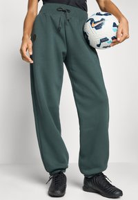 Grønne joggebukser med elastisk linning og snøring, med mansjetter ved ankelen. Kombinert med en svart fotball og svarte fotballevin.