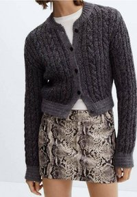 Mørkegrå kabelstrik cardigan med knaplukning, cropped design. Båret over en cremefarvet top og parret med en mønstret slangeskindsprint nederdel.